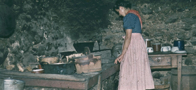 Historisches Foto in verblichenen Farben aus den fünfziger Jahren: Eine Frau an einem alten Herd. im Hintergrund ist eine alte graue Steinwand.