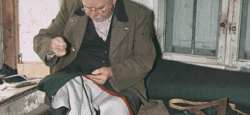 Historisches Foto in verblichenen Farben aus den fünfziger Jahren: Ein Mann sitz und näht mit den Händen an einer Tracht.